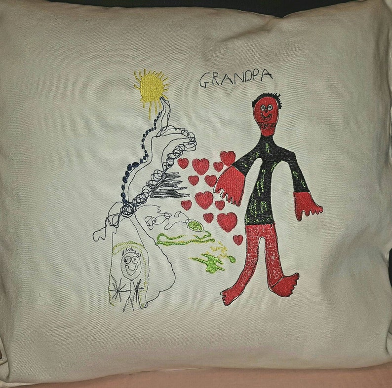 Peut inclure: Un oreiller blanc avec un motif brod&eacute; color&eacute;. Le motif repr&eacute;sente un personnage en b&acirc;ton avec des bras et des jambes rouges, un torse noir et un soleil jaune. Le texte "GRANDPA" est brod&eacute; en noir au-dessus de la figure. Il y a des c&oelig;urs rouges &eacute;parpill&eacute;s autour de la figure.