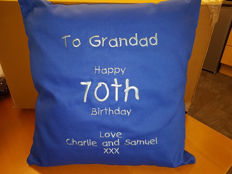 Peut inclure: Un coussin bleu avec une broderie blanche qui dit "To Grandad Happy 70th Birthday Love Charlie and Samuel XXX"