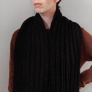 May include: A person wearing a black knitted scarf over a brown sweater. The scarf is thick and has a cable knit pattern.