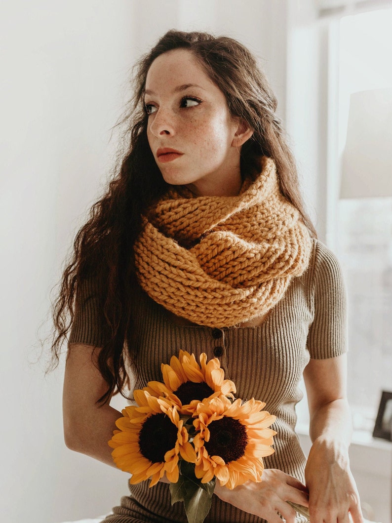 Pode incluir: Uma mulher com cabelo castanho comprido vestindo um su&eacute;ter de tric&ocirc; marrom e uma echarpe de tric&ocirc; amarela segura um buqu&ecirc; de girass&oacute;is.