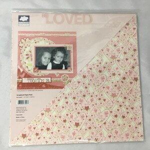 May include: A scrapbook paper pack with a pink and white heart pattern. The pack includes a sheet with a pink background and a photo frame with two children in it. The text "Be Loved" and "Happy Together" is printed on the paper.