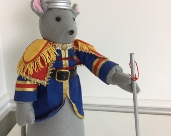May include: A gray mouse plush toy wearing a silver thimble hat and a blue, red, and gold uniform. The mouse is holding a silver stick with a red string attached to the top.