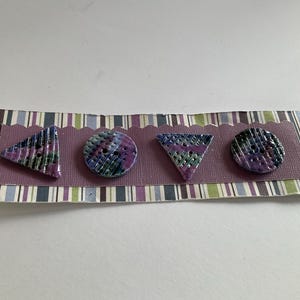 May include: Four decorative buttons in geometric shapes: triangle, circle, triangle, and circle. The buttons are purple, green, and blue with a textured pattern. They are attached to a purple card with a striped border.