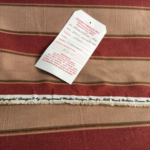May include: Striped fabric in shades of red, tan, and brown. The fabric has horizontal stripes and a tag attached with text. The bottom edge of the fabric is frayed. The tag reads "NRAVET FABRICS INC."