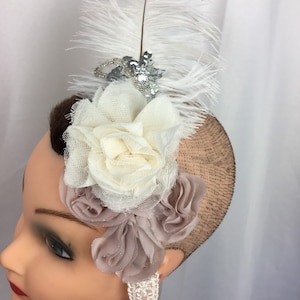 Pode incluir: Uma faixa de cabelo de penas brancas com uma flor branca grande e uma flor rosa menor. A faixa de cabelo é decorada com lantejoulas prateadas.