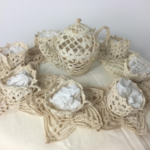 May include: A cream-colored, crocheted tea set with a teapot, cups, and a decorative doily. The teapot and cups have an openwork design. The cups are filled with white paper. The set is displayed on a white surface.