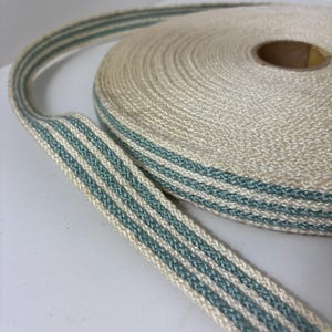 Peut inclure: Un rouleau de ruban crème avec trois rayures parallèles de couleur sarcelle. Le ruban est tissé serré et semble être fait d'une fibre naturelle. Le rouleau est partiellement déroulé, révélant le motif du ruban.