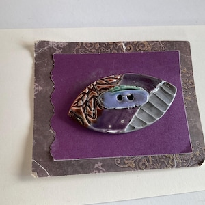 Puede incluir: Un botón ovalado con un diseño morado, marrón y gris. El botón tiene dos agujeros y está colocado sobre una tarjeta morada con un borde decorativo. La superficie del botón está dividida en secciones con diferentes texturas y colores.