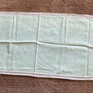 May include: A pale green oval-shaped table runner with a white lace trim. The runner is made of a soft fabric and has a subtle floral pattern. The lace trim adds a delicate touch to the overall design.