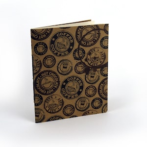 May include: A brown notebook with a coffee-themed design. The cover features repeating circular stamps with coffee cups, the words "Coffee," "Java," and "Hot Chocolate" in black ink. The notebook is open, revealing blank pages.