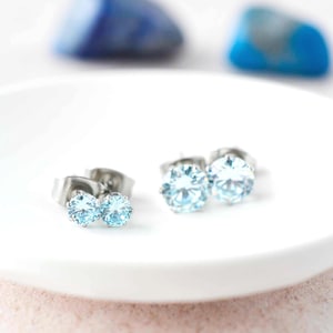 May include: A pair of stud earrings featuring round, light blue faceted stones set in silver-toned metal prongs. The earrings are displayed on a white surface, showcasing the clear, brilliant cut of the stones and the metallic post backings.