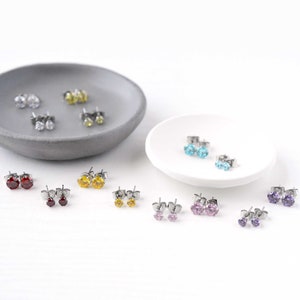 Peut inclure: Assortiment de boucles d'oreilles clous colorées avec des pierres précieuses claires, rouges, jaunes, bleues, roses et violettes. Les boucles d'oreilles sont présentées sur deux petits plats.