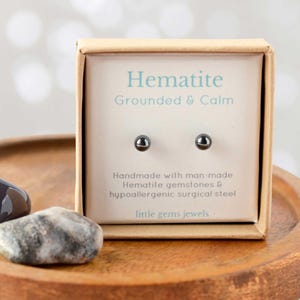 May include: A small box containing a pair of hematite stud earrings. The box is brown with white text that reads "Hematite Grounded & Calm Handmade with man-made Hematite gemstones & hypoallergenic surgical steel little gems jewels".