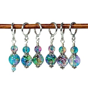 May include: Set of six colourful glass bead stitch markers with silver metal rings. The beads are iridescent and have a marbled pattern in shades of blue, green, and purple.