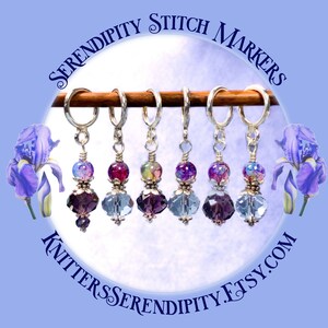 May include: Six iridescent glass bead stitch markers with silver rings and crystal accents. The markers are displayed on a wooden dowel rod. The text "Serendipity Stitch Markers" is displayed in a circle around the image.