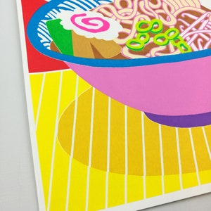 Pu&ograve; includere: Una ciotola rosa di ramen con noodles, cipolle verdi, una torta di pesce rosa e bianca e un uovo marrone. La ciotola &egrave; su una superficie a strisce gialle e bianche.
