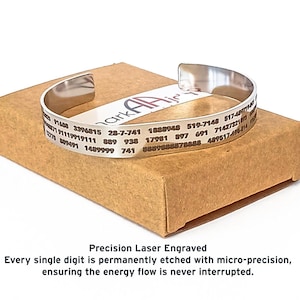 Pode incluir: Uma pulseira de punho prateada com números e texto gravados, repousando sobre uma caixa de cartão castanha. A pulseira tem um design simples e aberto. O texto na parte inferior diz "Precision Laser Engraved".