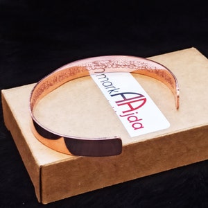 A copper bracelet with an ayatal Kursi engraved design. The bracelet is on a brown box