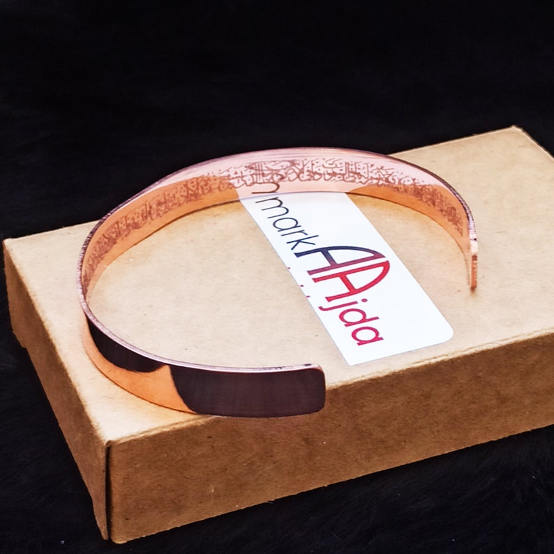 A copper bracelet with an ayatal Kursi engraved design. The bracelet is on a brown box