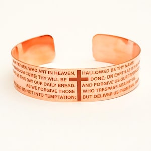 May include: Copper bracelet with the Lord's Prayer engraved in brown lettering. The bracelet features a small cross.