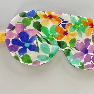 May include: A floral eye mask with a white background and a colourful pattern of flowers in shades of blue, purple, orange, and green. The mask has a yellow elastic band. The eye mask is designed to block out light.