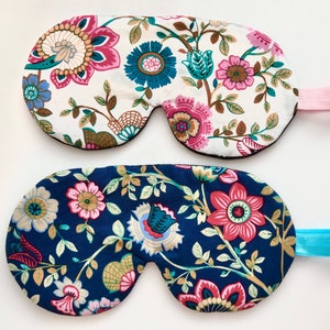 May include: Two floral eye masks. One is white with pink, teal, and blue flowers. The other is navy blue with similar floral patterns. Both have elastic bands for securing around the head. The eye masks are designed to block out light.