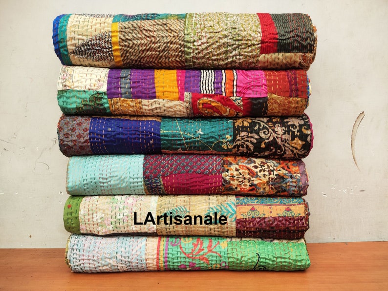 May include: A stack of colorful kantha quilts made from recycled saris. The quilts are layered in a variety of patterns and colors, including blue, green, purple, pink, and yellow. The quilts are hand-stitched and have a unique, vintage look.