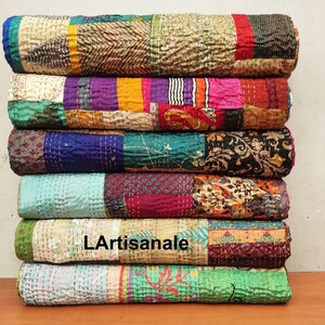 May include: A stack of six colorful kantha quilts, each with a different patchwork design. The quilts are made of recycled saris and are hand-stitched. The quilts are folded and stacked on a wooden surface. The text "L'Artisanale" is visible on one of the quilts.