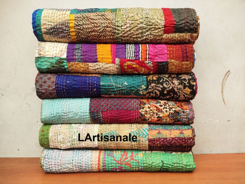 May include: A stack of six colorful kantha quilts, each with a different patchwork design. The quilts are made of recycled saris and are hand-stitched. The quilts are folded and stacked on a wooden surface. The text "L'Artisanale" is visible on one of the quilts.