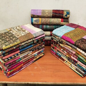 May include: A stack of colorful kantha quilts made from recycled saris. The quilts are made of various fabrics and patterns, including floral, paisley, and geometric designs. The quilts are arranged in two stacks on a wooden table.