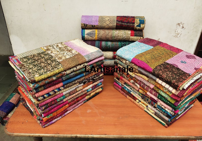 May include: A stack of colorful kantha quilts made from recycled saris. The quilts are made of various fabrics and patterns, including floral, paisley, and geometric designs. The quilts are arranged in two stacks on a wooden table.