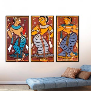 May include: Three framed paintings of women in traditional Indian clothing. The women are depicted in a colorful, stylized manner with bright yellow, blue, and red accents. The paintings are set against a white wall.