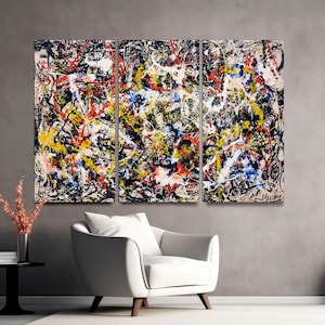 May include: Abstract expressionist artwork in a triptych format. The canvas features a dynamic composition of black, white, yellow, red, and blue paint splatters and lines. The artwork is displayed above a white armchair and a black side table.