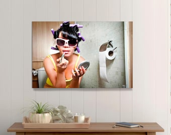 May include: A woman in a yellow top and sunglasses applies lipstick while looking in a compact mirror. She has purple curlers in her hair. A roll of toilet paper hangs on the wall behind her.