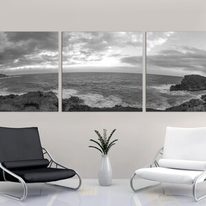 May include: A black and white panoramic photograph of a rocky coastline with crashing waves and a cloudy sky. The photograph is split into three panels and is displayed on a wall above two modern chairs.