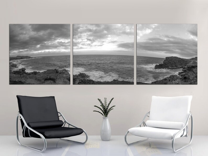 May include: A black and white panoramic photograph of a rocky coastline with crashing waves and a cloudy sky. The photograph is split into three panels and is displayed on a wall above two modern chairs.