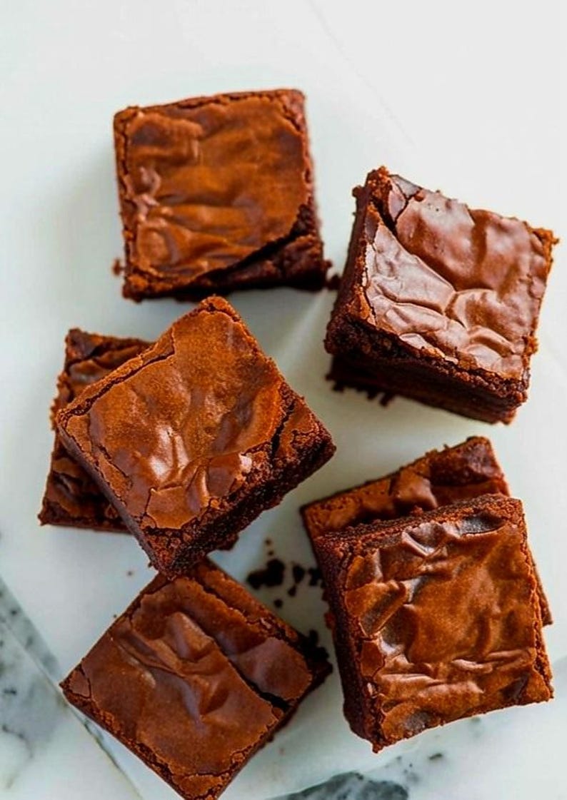 Pu&ograve; includere: Vista dall'alto di sei brownies quadrati con un ricco colore marrone scuro. I brownies hanno una superficie leggermente screpolata e strutturata, che suggerisce un interno morbido. Disposti su una superficie bianca.