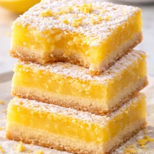 May include: Three lemon bars stacked on top of each other. The bars have a buttery crust, a bright yellow filling, and are dusted with powdered sugar and lemon zest. The top bar has a bite taken out of it.
