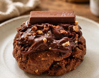 May include: A round chocolate cookie topped with a thick layer of dark chocolate frosting, scattered cookie crumbles, and a single rectangular chocolate wafer bar with the partial text Kit visible on the surface, served on a light speckled plate.