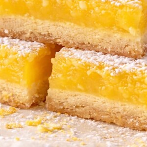 May include: Close-up of lemon bars dusted with powdered sugar. The bars have a buttery, golden crust and a bright yellow, citrusy filling. The dessert is arranged on a white plate.