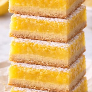 Pu&ograve; includere: Una pila di cinque barrette al limone. Ogni barretta ha una base burrosa marrone dorato sormontata da un ripieno giallo brillante e agrumato. Le barrette sono spolverate con zucchero a velo e disposte su una superficie bianca.