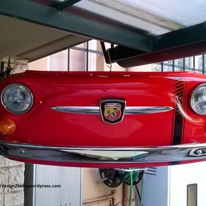 Op de afbeelding: De voorkant van een rode Fiat 500 met een zwart-witte streep aan de zijkant. De auto hangt aan het plafond.
