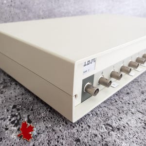 Może przedstawiać: Białe pudełko z etykietą LEONI i wieloma czarnymi złączami po bokach. Pudełko stoi na szarej, teksturowanej powierzchni.