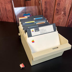 Puede incluir: Una caja de almacenamiento de plástico beige con una tapa transparente, llena de disquetes Apple II vintage. El disco superior está etiquetado como "MousePaint Drawing Program for the Apple II and Mouse II".
