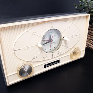 May include: Cream-colored vintage alarm clock with a black top and a retro design. The clock face is oval with black hands and white numerals. Features two knobs and the name "Fleetwood".