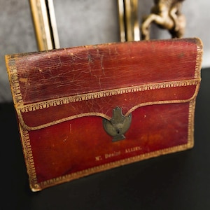Puede incluir: Un maletín de documentos vintage de cuero rojo con un ribete dorado decorativo y un cierre metálico. El maletín presenta desgaste visible, lo que sugiere antigüedad. El texto "M. Desire Allier" es visible.