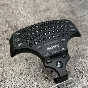 Könnte beinhalten: Eine schwarze Sony PlayStation-Tastatur mit kreisförmigem Layout. Die Tastatur verfügt über eine Vielzahl von Tasten, darunter Buchstaben, Zahlen und Symbole. Die Tastatur verfügt außerdem über ein Richtungspad, eine Select-Taste und eine Start-Taste. Die Tastatur befindet sich in einem durchsichtigen Plastikständer.