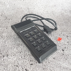 Puede incluir: Un Video Touch Pad negro con una cuadrícula de botones cuadrados y un cable. El texto "VIDEO TOUCH PAD" es visible en la parte superior. Una pequeña hoja de arce roja está al lado de la almohadilla.