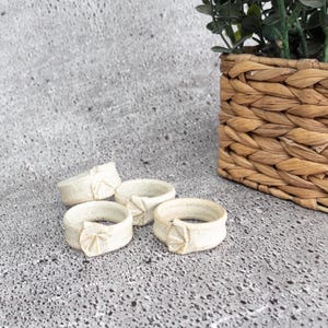 May include: Four off-white ceramic napkin rings with a leaf design. The rings are arranged on a speckled grey surface, with a woven basket containing greenery in the background. The napkin rings are a decorative element for table settings.
