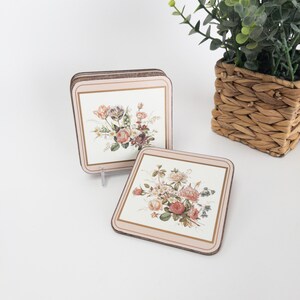 Op de afbeelding: Set van twee vierkante onderzetters met bloemmotieven. Elke onderzetter heeft een crèmekleurige achtergrond met een boeket van roze, perzik en paarse bloemen. De onderzetters hebben een lichtroze rand met een gouden rand.